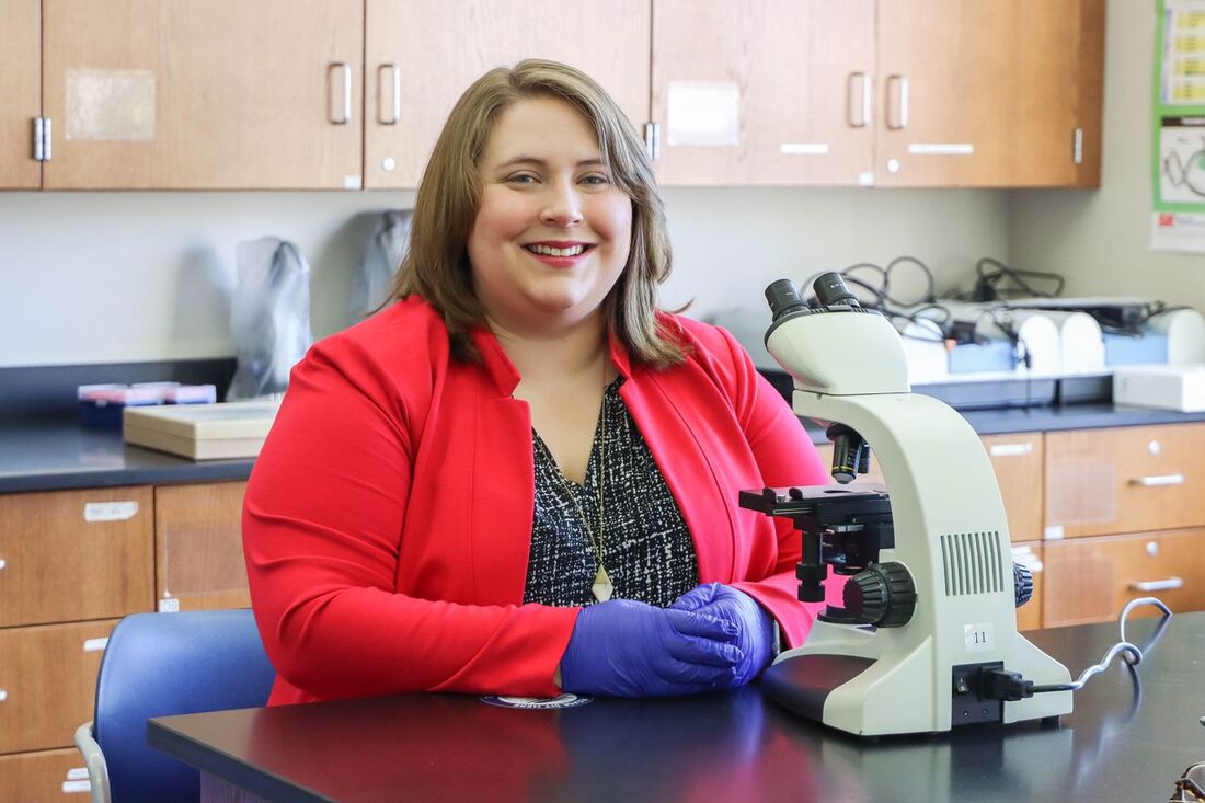 Dr. Tokash-Peters in a red blazer sitting behind a microscope smiling and wearing purple nitrile gloves.